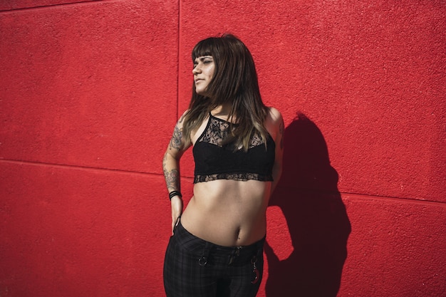 Jeune femme de race blanche attrayante avec des tatouages debout contre un mur rouge