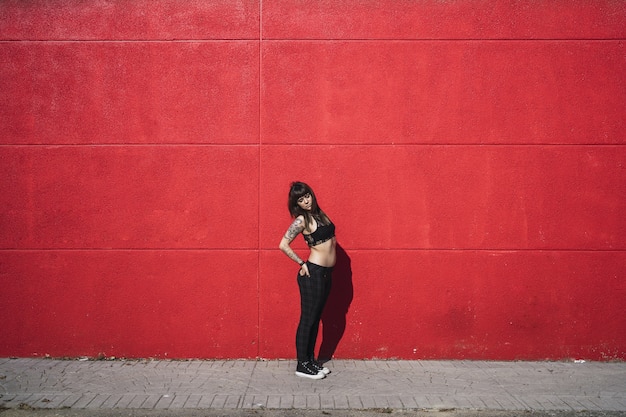 Jeune femme de race blanche attrayante avec des tatouages debout contre un mur rouge