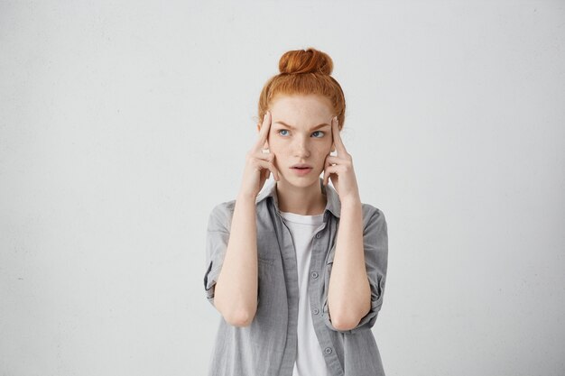 Jeune femme de race blanche rousse sérieuse tenant les doigts sur ses tempes et regardant sur le côté avec une expression concentrée et concentrée, comme si elle essayait de se souvenir de quelque chose d'important