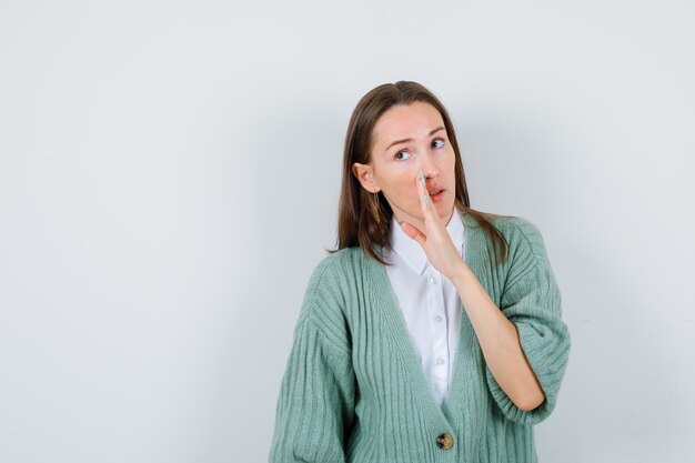 Jeune femme racontant le secret derrière la main en chemise, cardigan et semblant prudente. vue de face.