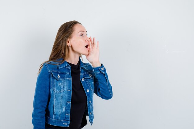 Jeune femme racontant le secret avec la main près de la bouche en blouse, vue de face.