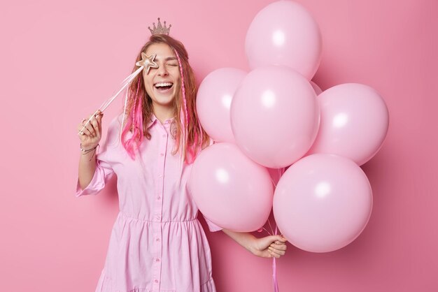 Une jeune femme ravie et heureuse pose avec un tas de ballons gonflés et une baguette magique s'amuse sur des idiots de fête autour d'isolé sur fond rose profite des vacances Personnes et concept de célébration