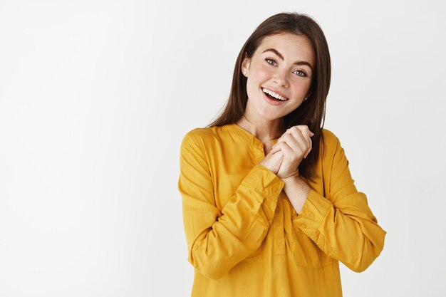 Jeune femme ravie et heureuse regardant avec admiration et joie, serrer les mains ensemble reconnaissante pour le cadeau, debout touchée sur un mur blanc.