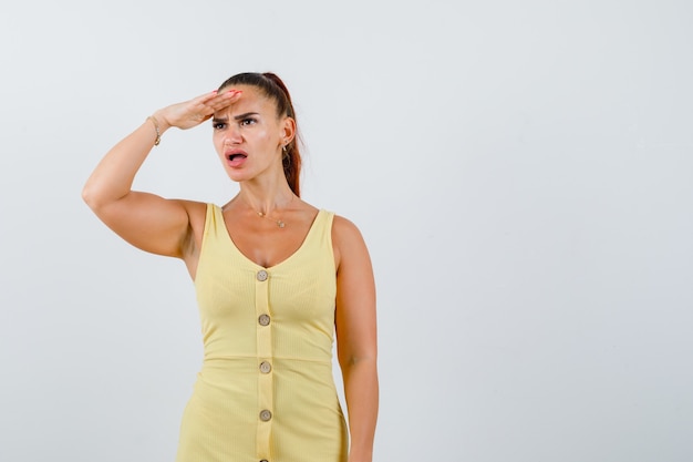 Jeune Femme à La Recherche De Loin Avec La Main Sur La Tête En Robe Jaune Et à La Frustration. Vue De Face.