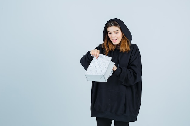 Jeune femme à la recherche en paquet dans un sweat à capuche surdimensionné, un pantalon et à la surprise. vue de face.