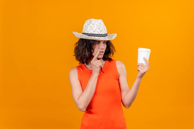 Une jeune femme réfléchie aux cheveux courts dans une chemise orange portant un chapeau de soleil à la tasse en plastique