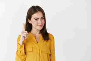 Photo gratuite jeune femme réfléchie montrant une publicité, pointant le doigt vers le haut et souriant à l'avant, donnant un regard conscient, debout sur un mur blanc