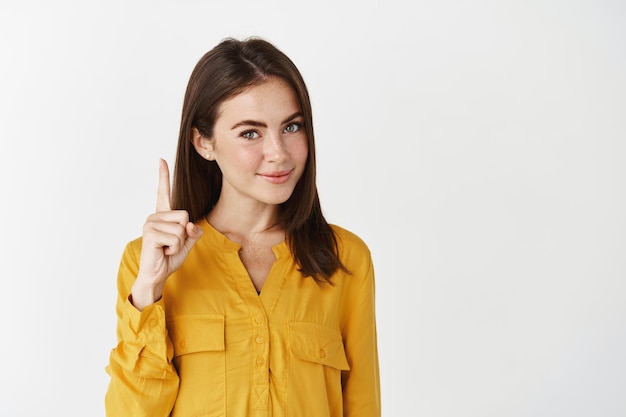 Jeune femme réfléchie montrant une publicité, pointant le doigt vers le haut et souriant à l'avant, donnant un regard conscient, debout sur un mur blanc