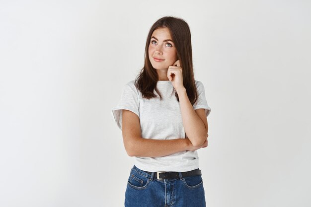 Jeune femme réfléchie prenant sa décision, regardant le coin supérieur gauche et souriant hésitant, debout sur un mur blanc.