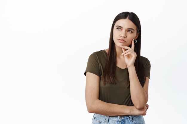 Jeune femme réfléchie regardant le coin supérieur gauche avec un visage sérieux et louche, besoin de réfléchir, faire des suppositions, debout pensive contre le mur blanc