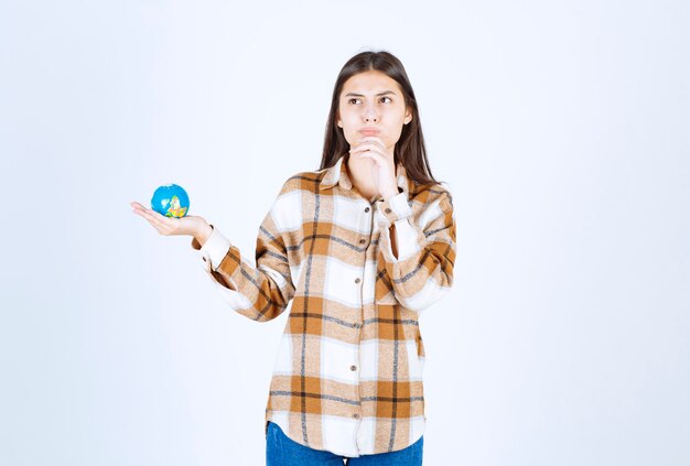 jeune femme réfléchie regardant un petit globe sur un mur blanc.