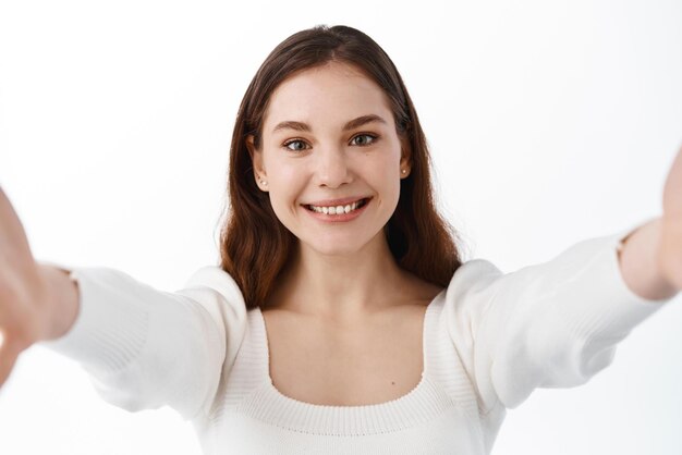 Jeune femme regardant la caméra du smartphone étirer les mains pour tenir le gadget pendant l'appel vidéo en prenant selfie sur un téléphone portable ou une tablette numérique debout sur fond blanc