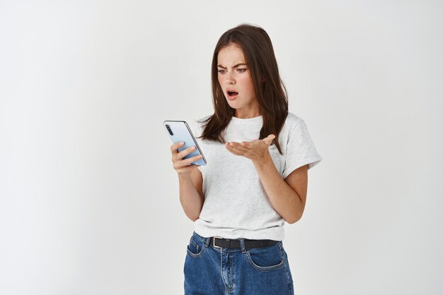 Jeune femme regardant en colère et confuse devant l'écran du smartphone se plaignant d'une application cassée debout sur fond blanc