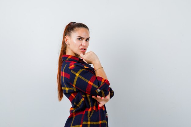 Jeune femme regardant par-dessus l'épaule tout en posant en haut court, chemise à carreaux, pantalon et charmante. vue de face.