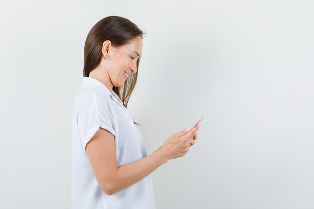 Jeune femme regardant son téléphone tout en souriant en chemisier blanc et à la recherche de bonheur.