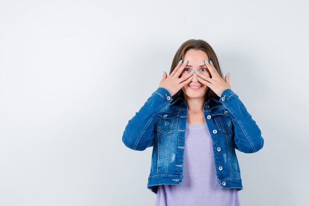 Jeune femme regardant à travers les doigts en t-shirt, veste et semblant joyeuse. vue de face.