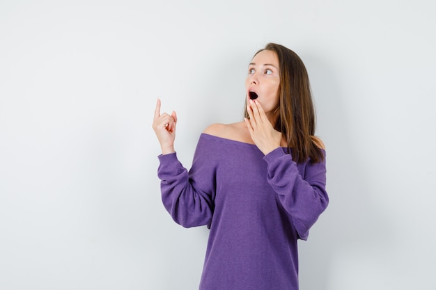 Jeune femme regardant vers le haut avec le doigt en chemise violette et à la surprise, vue de face.