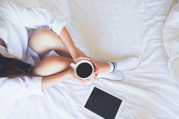 Une jeune femme relaxante apprécie son café en assise dans son lit.