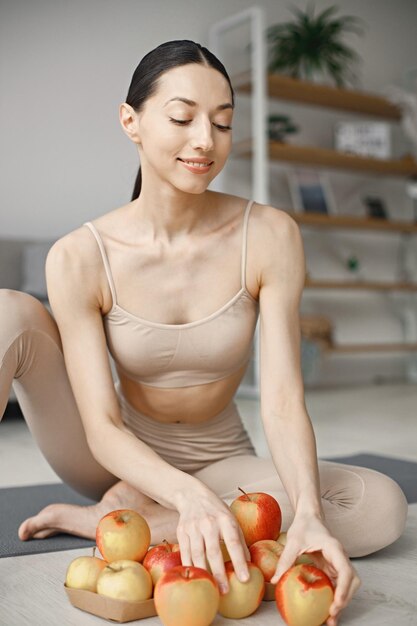 Jeune femme de remise en forme assise sur un tapis de yoga à la maison et tenant des pommes fraîches