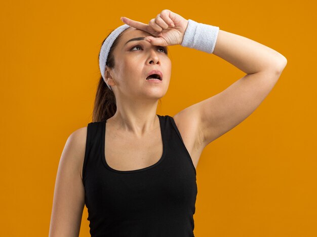 Jeune femme de remise en forme avec bandeau et brassards levant la main sur la tête fatiguée et surmenée debout sur un mur orange