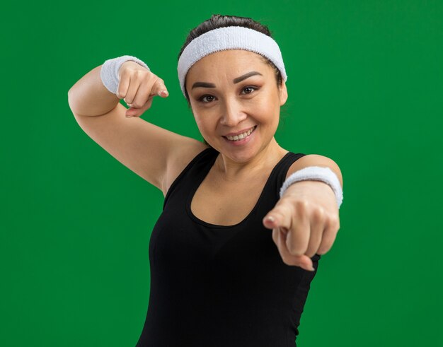 Jeune femme de remise en forme avec bandeau et brassards souriant avec un visage heureux pointant avec l'index debout sur un mur vert