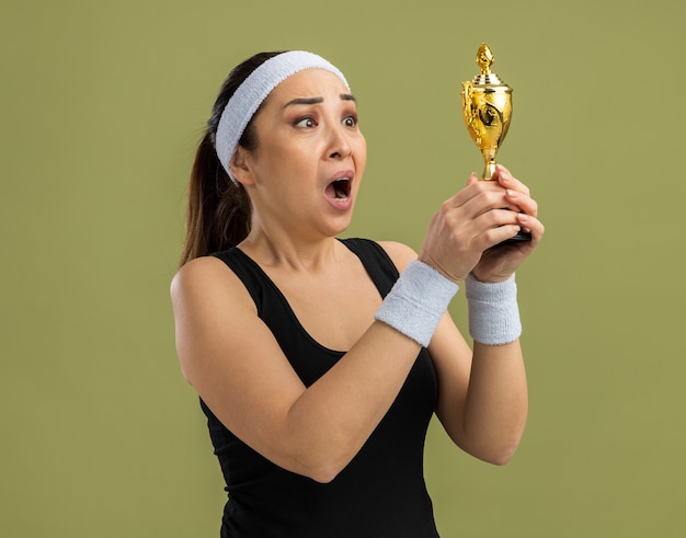 Jeune femme de remise en forme avec bandeau et brassards tenant un trophée en le regardant avec une expression déçue debout sur un mur vert