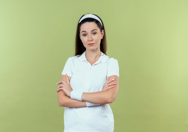 Jeune femme de remise en forme en bandeau avec une expression confiante avec les bras croisés debout sur un mur léger