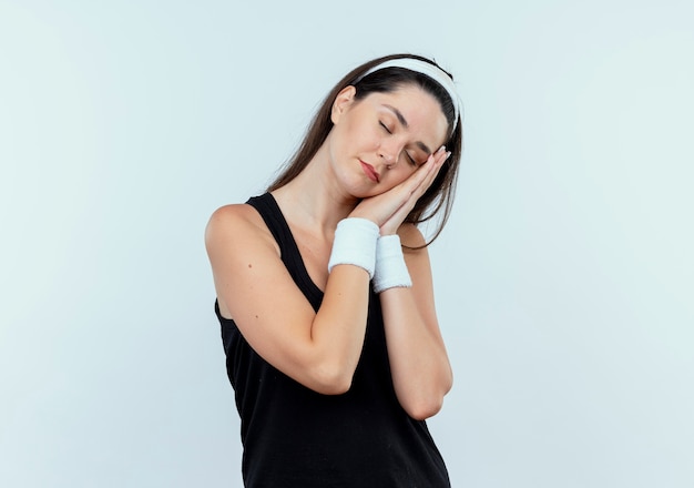 Jeune femme de remise en forme en bandeau faisant le geste de sommeil avec les paumes de la tête penchée sur les paumes avec les yeux fermés debout sur fond blanc