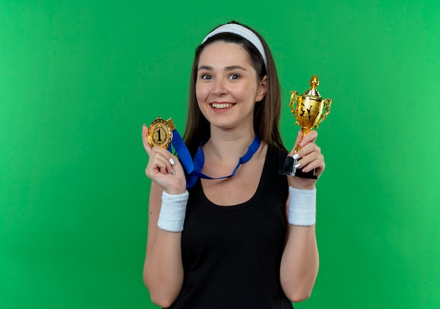 Jeune femme de remise en forme en bandeau avec médaille d'or autour de son cou tenant le trophée regardant la caméra en souriant avec un visage heureux debout sur fond vert