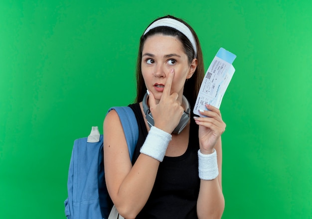 Photo gratuite jeune femme de remise en forme en bandeau avec sac à dos tenant des billets d'avion pointant avec le doigt à son œil debout sur fond vert