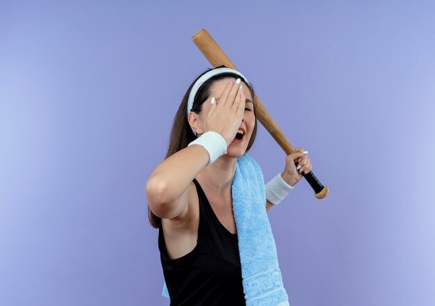 Jeune femme de remise en forme en bandeau avec une serviette sur son épaule tenant une batte de baseball couvrant un œil avec une main souriant debout sur fond bleu