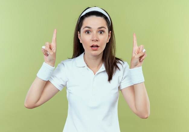 Jeune femme de remise en forme en bandeau surpris montrant les index ayant une nouvelle idée debout sur un mur léger