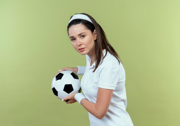 Jeune femme de remise en forme en bandeau tenant un ballon de football avec un visage sérieux debout sur un mur léger