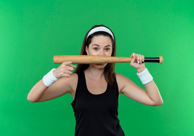 Jeune femme de remise en forme en bandeau tenant une batte de baseball pointant avec le doigt souriant debout sur un mur vert
