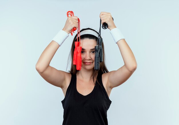 Jeune femme de remise en forme en bandeau tenant la corde à sauter regardant la caméra avec le sourire sur le visage debout sur fond blanc