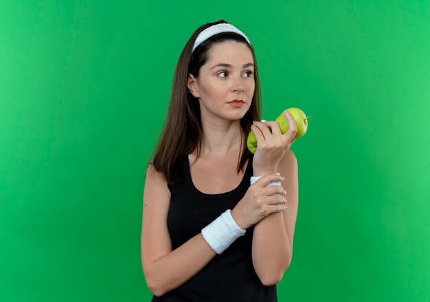 Jeune femme de remise en forme en bandeau tenant des pommes vertes à côté avec un visage sérieux debout sur fond vert