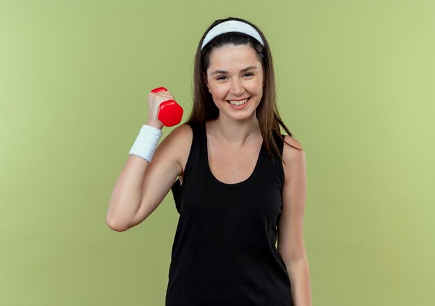 Jeune femme de remise en forme en bandeau travaillant avec haltère souriant avec visage heureux debout sur fond clair