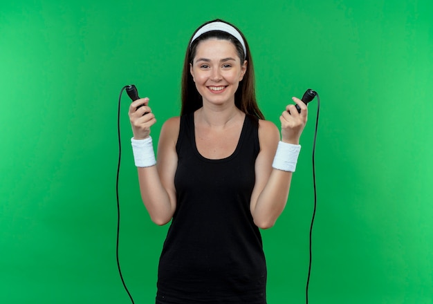 Jeune femme de remise en forme dans le bandeau tenant la corde à sauter souriant joyeusement va sauter debout sur le mur vert