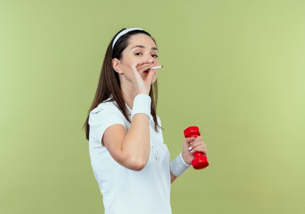 Jeune femme de remise en forme dans le bandeau travaillant avec haltère et fumer une cigarette debout sur un mur léger