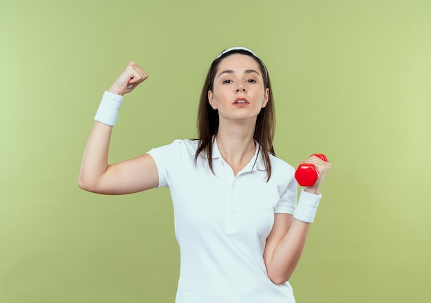 Jeune femme de remise en forme dans le bandeau travaillant avec haltère montrant les biceps avec un visage sérieux debout sur un mur léger