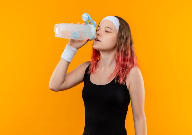 Jeune femme de remise en forme dans l'eau potable de sportswear avec les yeux fermés debout sur le mur orange