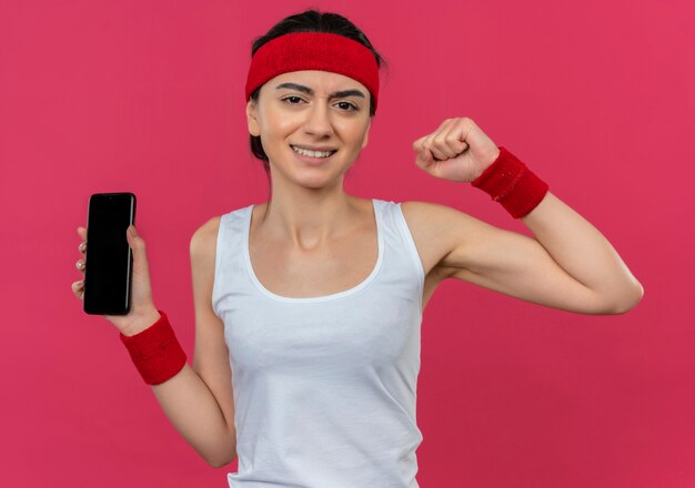 Jeune femme de remise en forme en tenue de sport avec bandeau montrant le poing de smartphone avec une expression agressive debout sur un mur rose