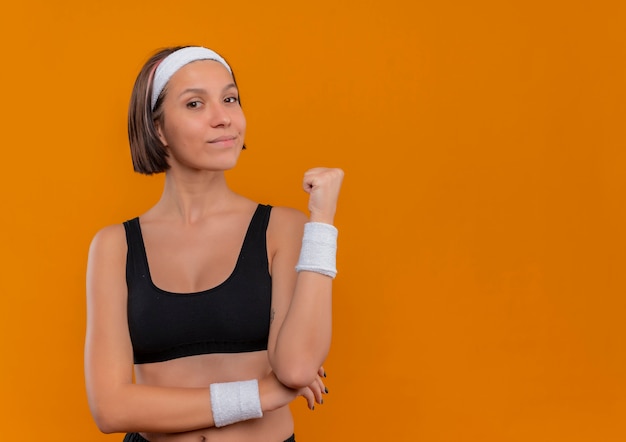 Jeune femme de remise en forme en tenue de sport avec bandeau, pointant vers l'arrière à la confiance debout sur le mur orange