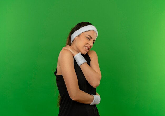 Jeune femme de remise en forme en tenue de sport avec bandeau à la recherche de mal en touchant son épaule ayant de la douleur debout sur le mur vert