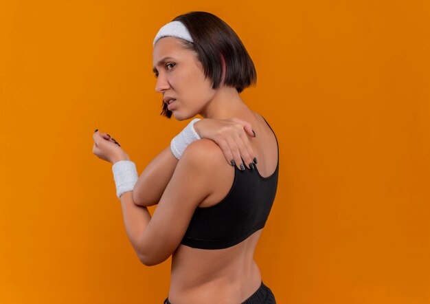 Jeune femme de remise en forme en tenue de sport avec bandeau à la recherche de mal en touchant son épaule sensation de douleur debout sur un mur orange