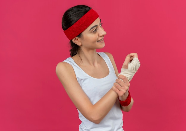 Jeune femme de remise en forme en tenue de sport avec bandeau à la recherche de mal en touchant son poignet bandé smiling positive debout sur le mur rose