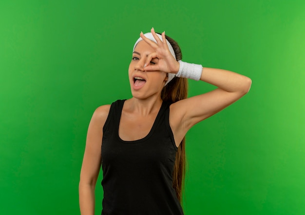 Jeune femme de remise en forme en tenue de sport avec bandeau souriant joyeusement faisant signe ok regardant à travers ce panneau debout sur mur vert