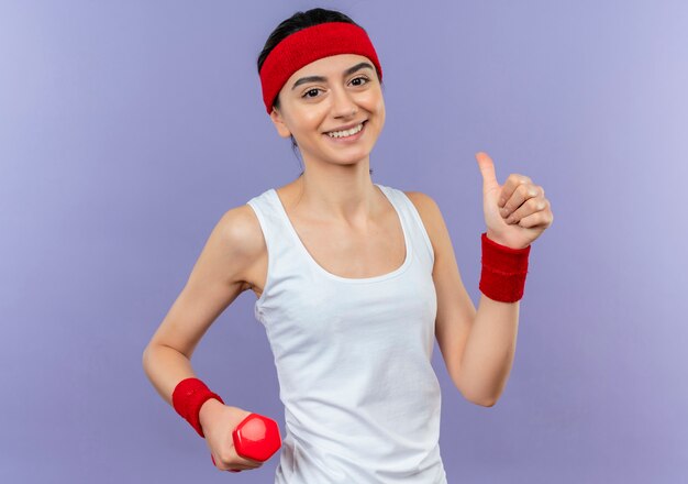 Jeune femme de remise en forme en tenue de sport avec bandeau souriant joyeusement montrant les pouces vers le haut debout sur le mur violet