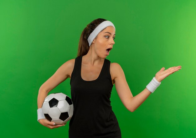 Jeune femme de remise en forme en tenue de sport avec bandeau tenant un ballon de football pointant avec le bras de sa main sur le côté à la surprise debout sur le mur vert