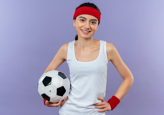 Jeune femme de remise en forme en tenue de sport avec bandeau tenant un ballon de football souriant confiant heureux et positif debout sur mur violet